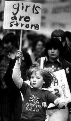 Black and white photograph of young girl marching with placard which says 'girls are strong'